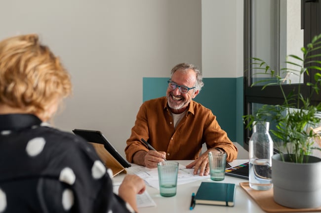 Harmaahiuksinen mies pöydän äärellä tarkastamassa asiakirjoja ja hymyilemässä katsoen kuvan ulkopuolella olevaa henkilöä.