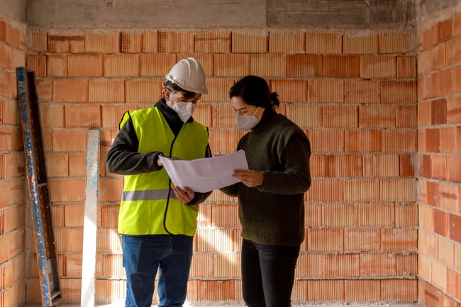 Keskellä kuvassa mies ja nainen työmaalla tarkastamassa suunnittelupapereita. Miehellä on työmaa-asustus päällä.